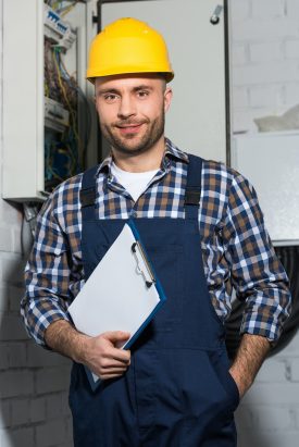 Electrician holding clipboard and smiling by electrical box